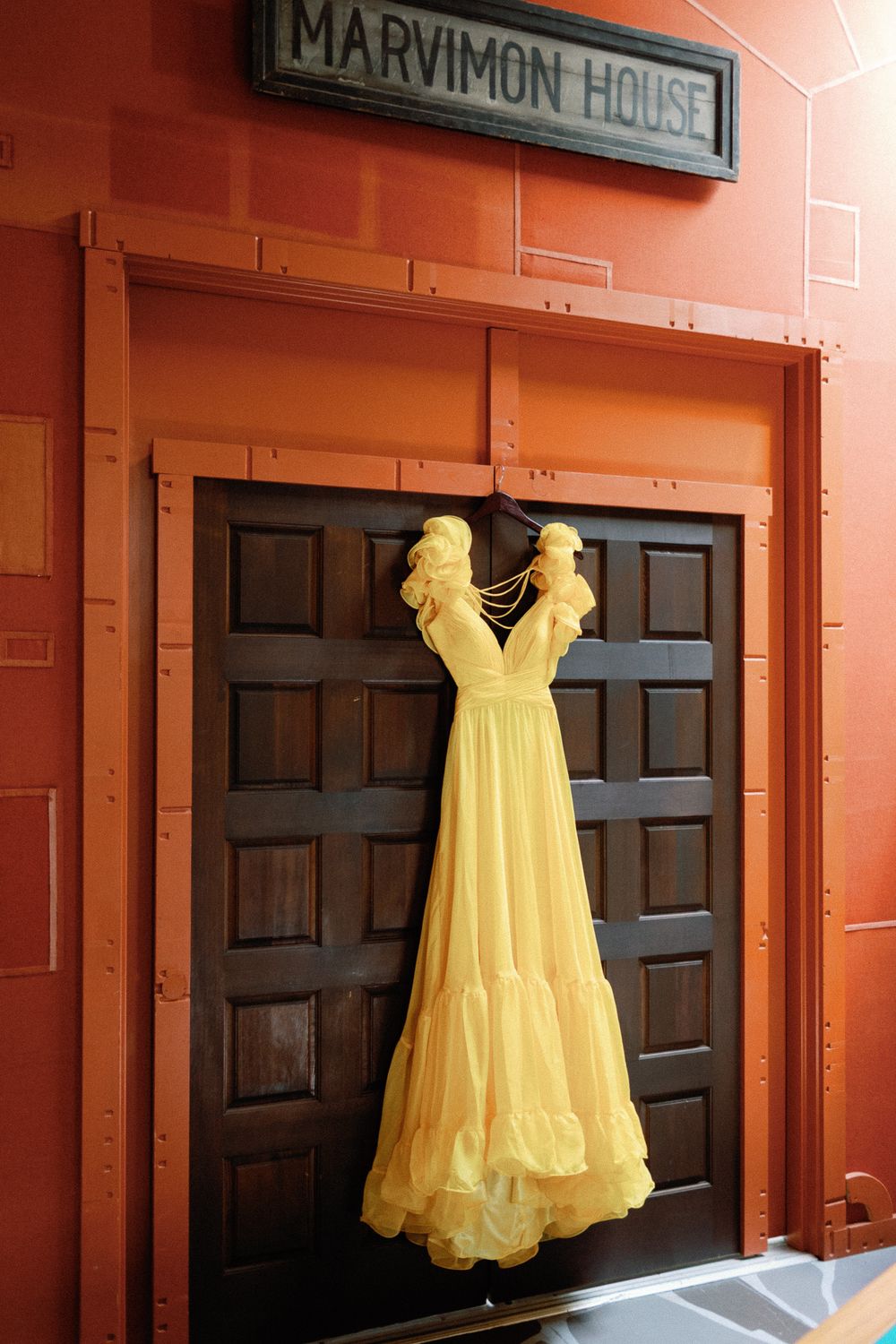 Yellow wedding dress hanging on dark wooden doors at Marvimon House with orange walls.