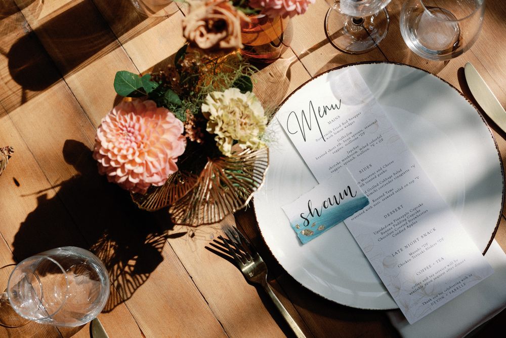 Elegant wedding place setting with menu cards on a silver plate, gold flatware, and pink floral centerpiece on a wood table.