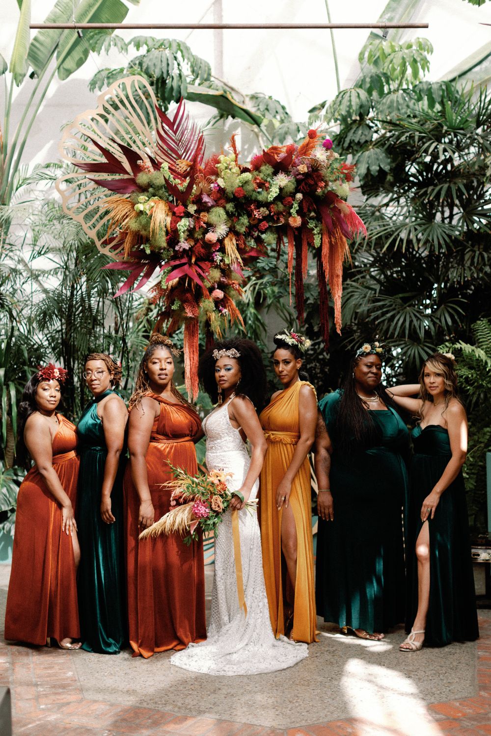 Bride in white lace gown with bridesmaids in jewel-toned velvet dresses posing in a tropical greenhouse setting.
