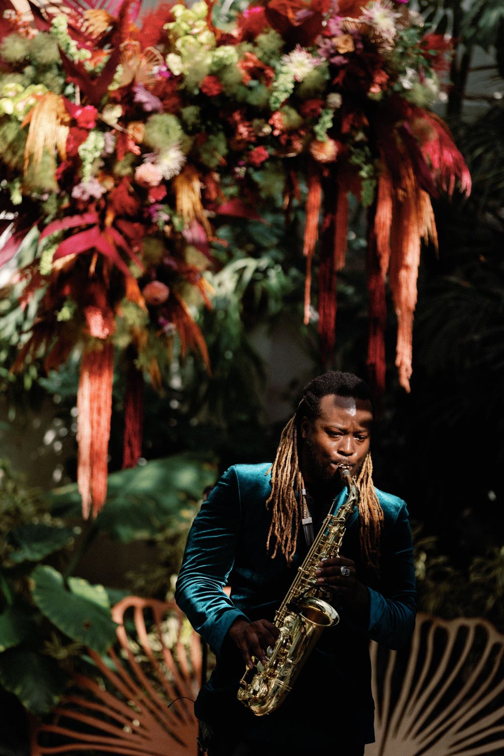 Saxophonist in teal velvet suit performing beneath a vibrant tropical floral arrangement in a lush greenhouse.