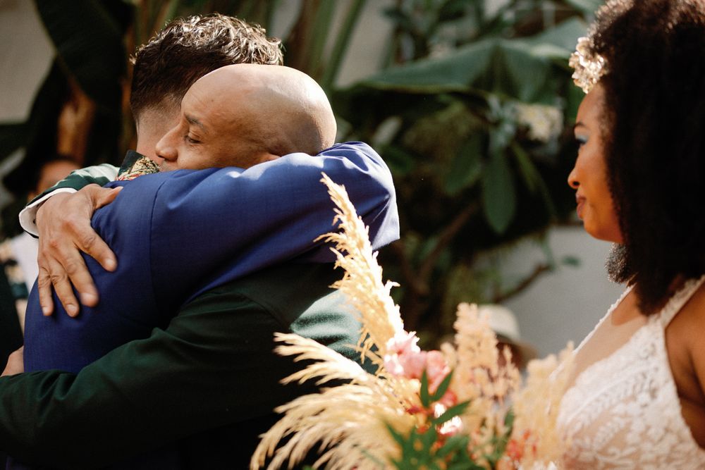 Emotional wedding ceremony moment as groom shares a heartfelt embrace with a guest while bride looks on warmly.