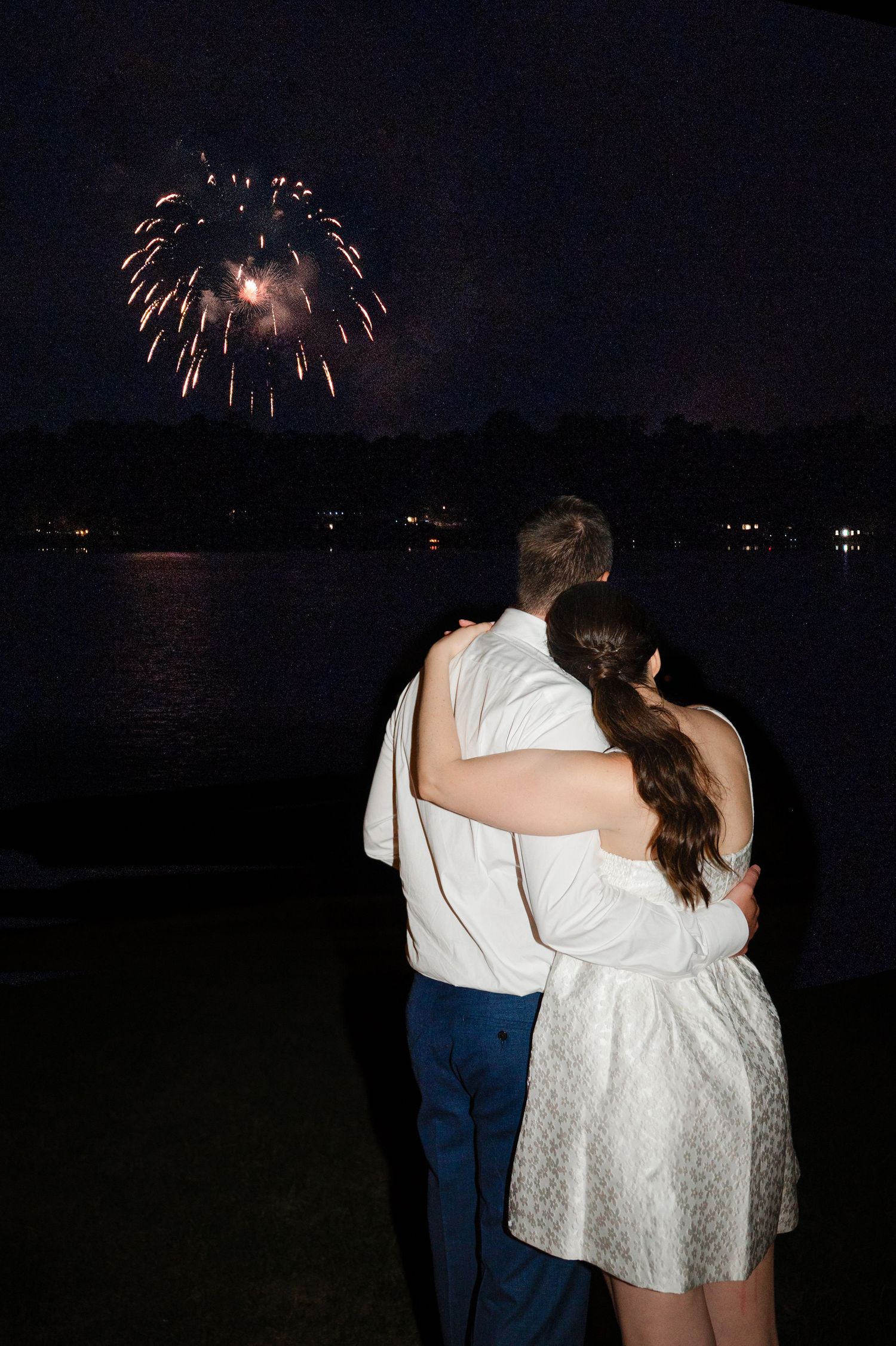Newlyweds watch colorful fireworks explode against the night sky over a lake, creating reflections on the water on their wedding night.