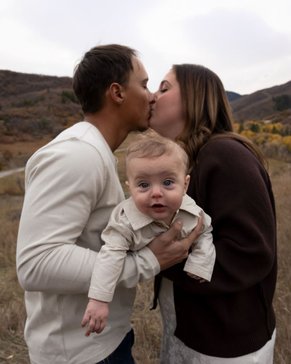A couple shares a romantic kiss while holding a baby outdoors in an autumn mountain landscape.