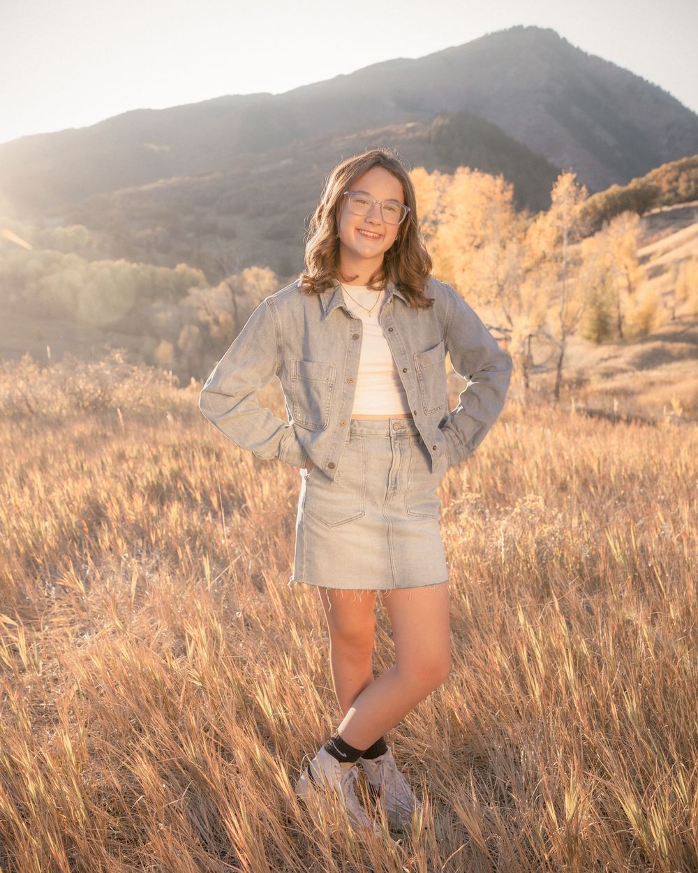 A person in a gray jacket poses in a golden field at sunset with mountains in the background.