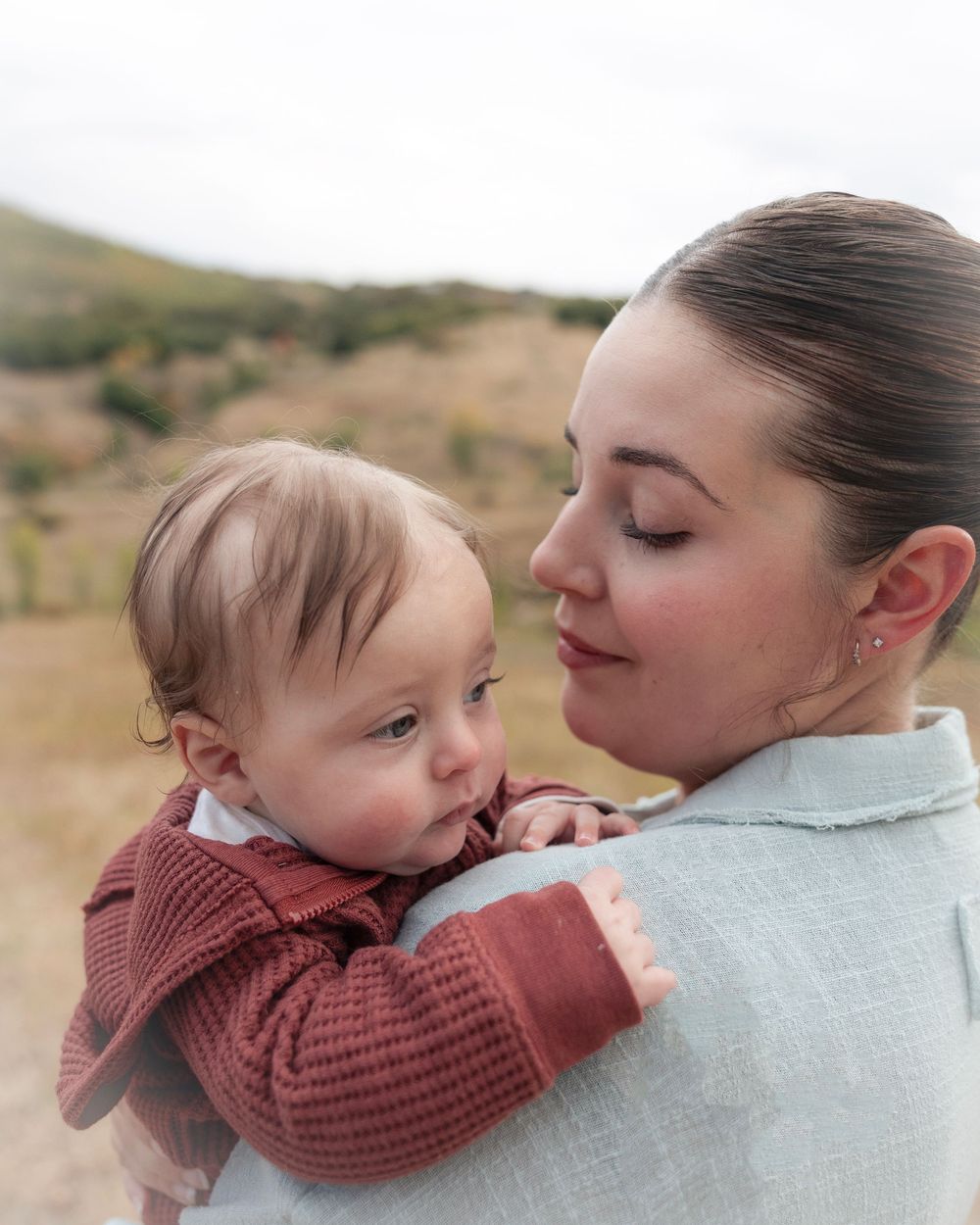 A tender moment captured outdoors as a parent holds their baby wearing a burgundy sweater against a natural landscape backdrop.