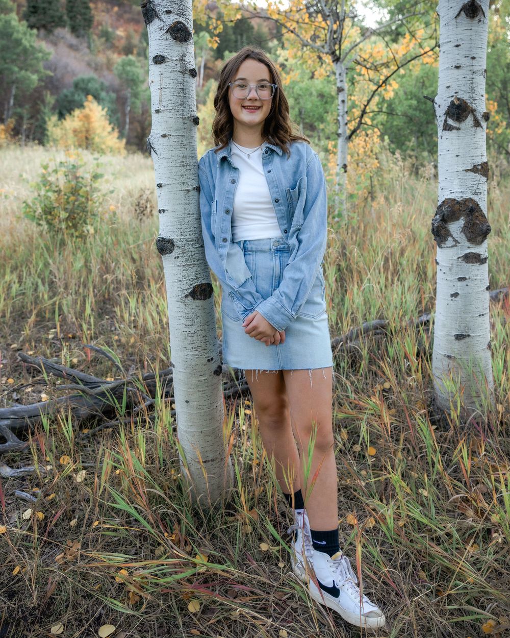 A person poses among aspen trees wearing a denim jacket and white sneakers during autumn.