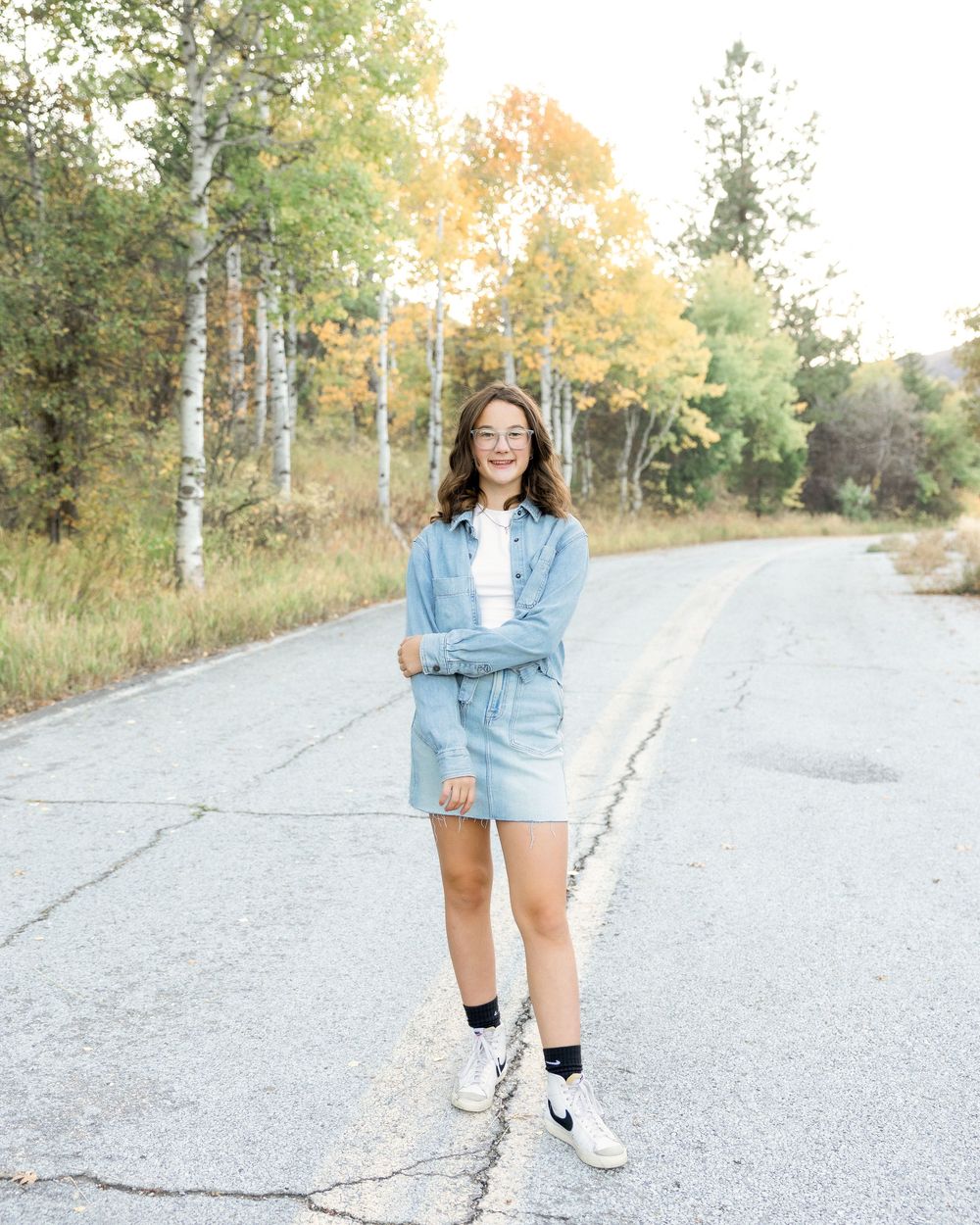 A person in a denim jacket and shorts poses casually on a gravel path surrounded by autumn trees.