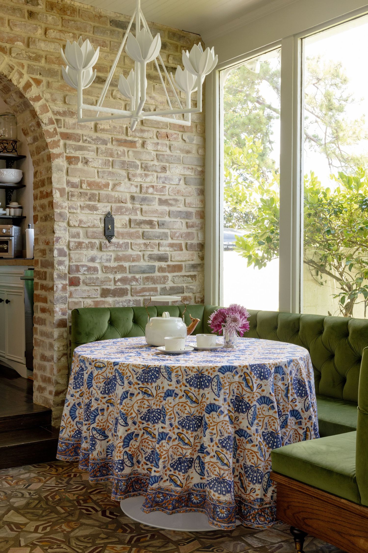 Cozy dining nook with exposed brick wall, green velvet banquette, floral tablecloth and white tulip chandelier.