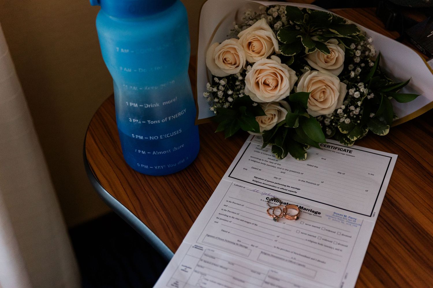Pastel peach roses and a blue bottle sit on a wooden table beside paperwork.