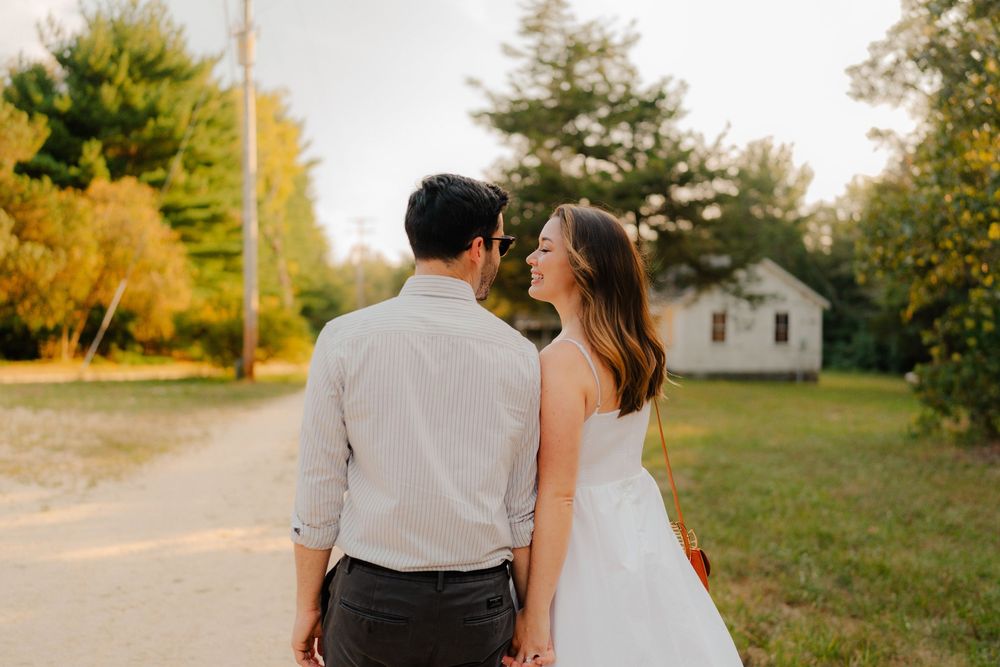 A couple in wedding attire walks together down a country road at sunset with autumn trees in the background.