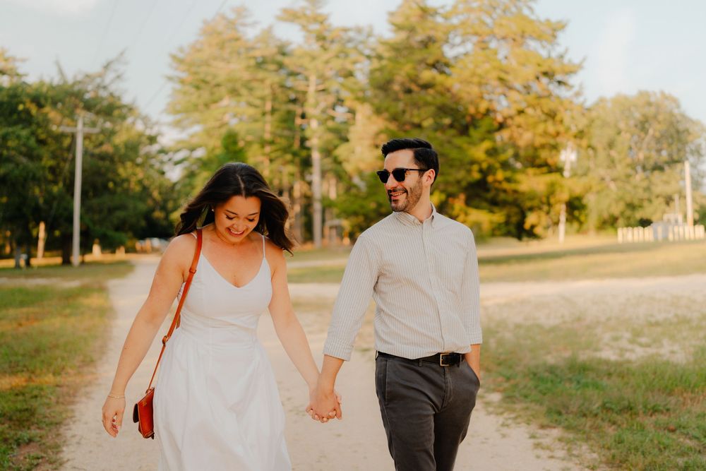 Couple walking together and holding hands on a dirt path during golden hour with autumn trees in background.