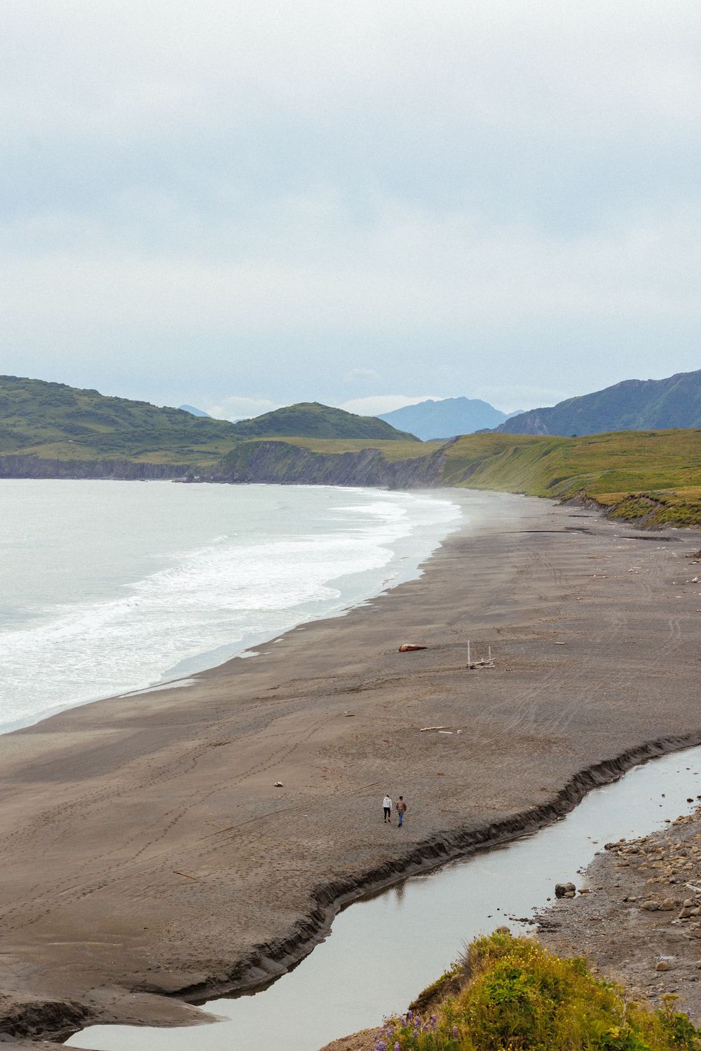 Scenic view of a long black sand beach with mountains in the distance and a small stream cutting through the sand.