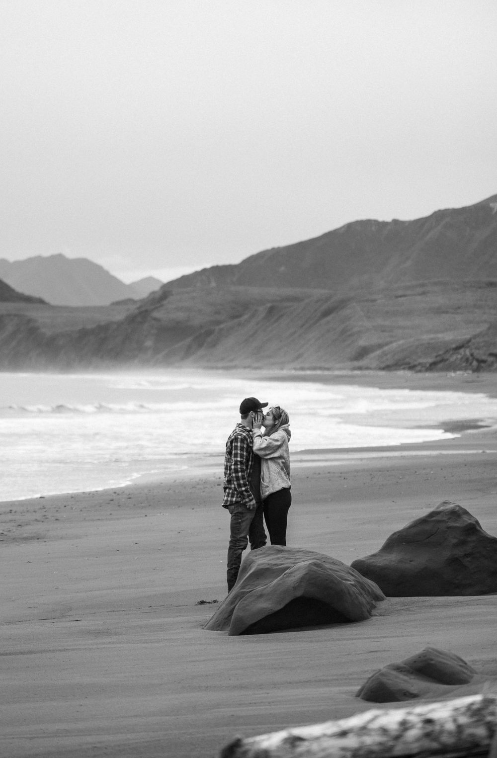 Two people embrace while standing on a misty beach with mountains and rocks in the background.