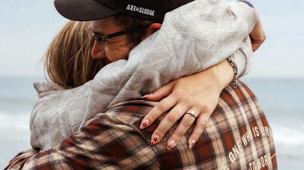 Two people share a warm embrace on a beach wearing casual clothing and a cap.