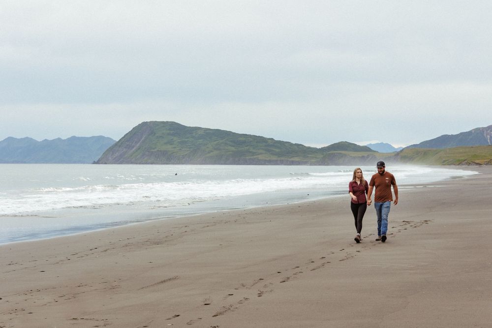 A couple walks hand in hand along a misty Pacific Northwest beach with mountains in the background.