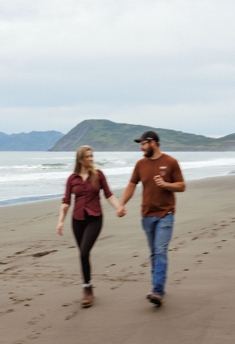 Couple taking a romantic stroll on a sandy beach with coastal mountains and waves visible.