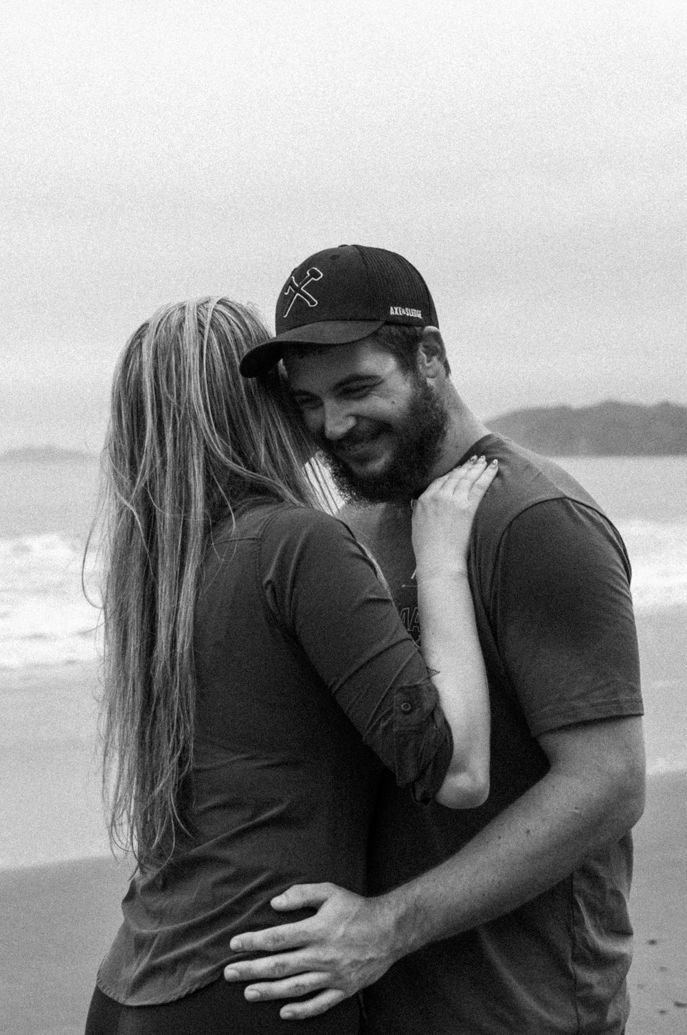 Romantic black and white photo of a couple embracing on a beach with ocean waves in background.