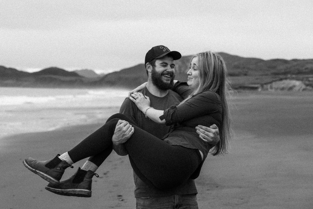 A romantic couple embracing and dancing on a scenic beach with mountains in the background.
