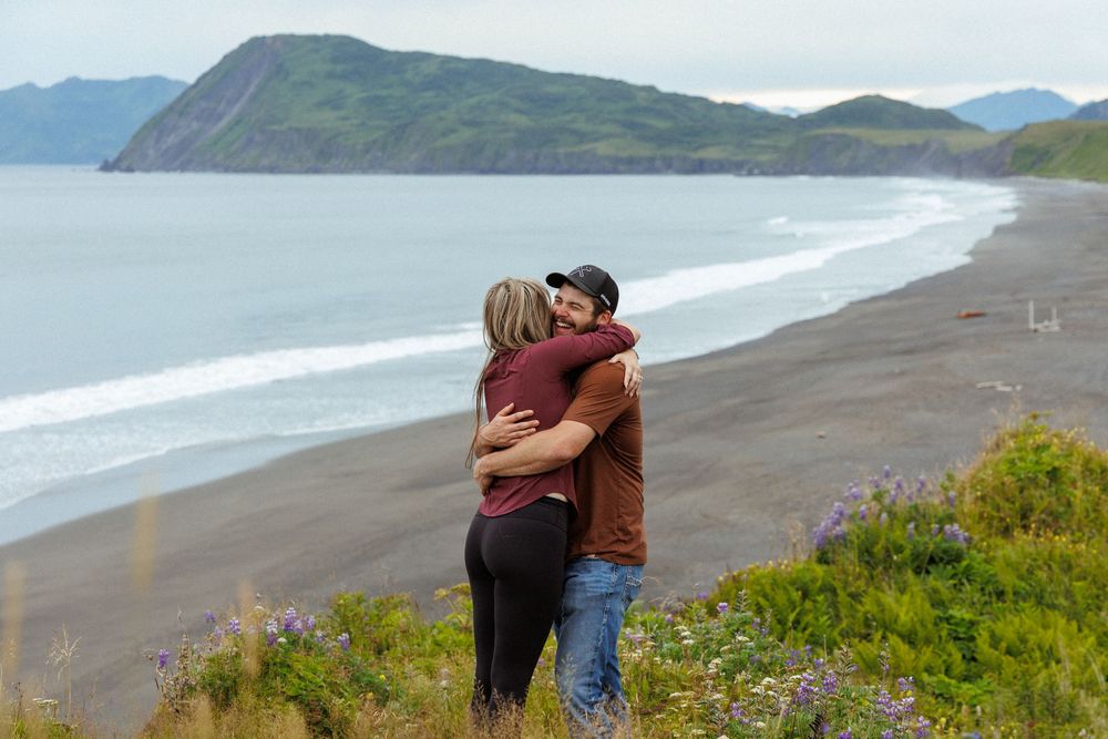 Couple embracing on cliffside overlooking dramatic coastal landscape and beach below.