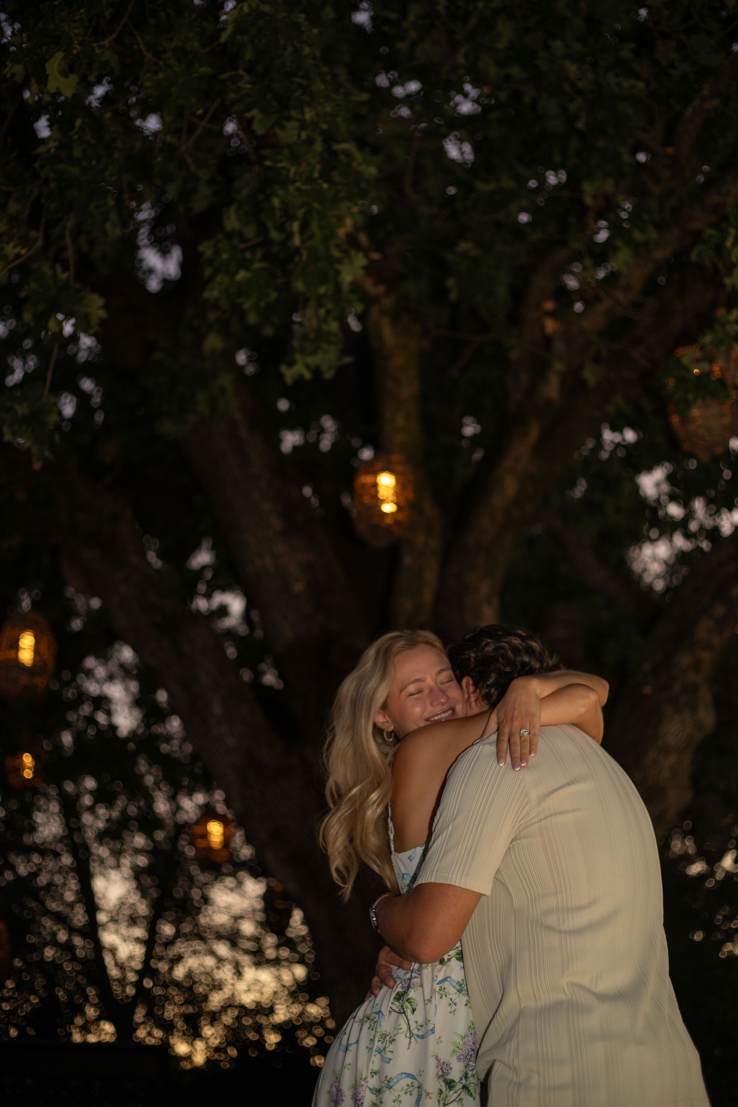 A romantic evening moment between two people is captured under trees with glowing lanterns at sunset.