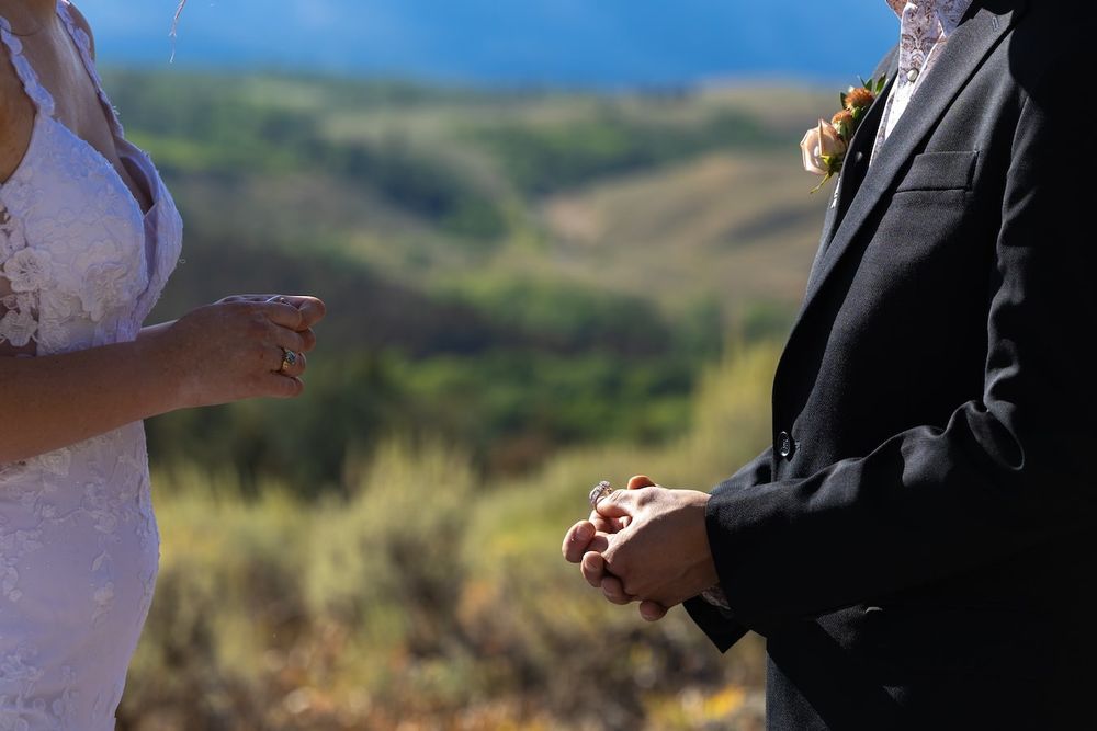 Two people exchange rings in front of guests during an outdoor wedding ceremony with a scenic mountain backdrop.