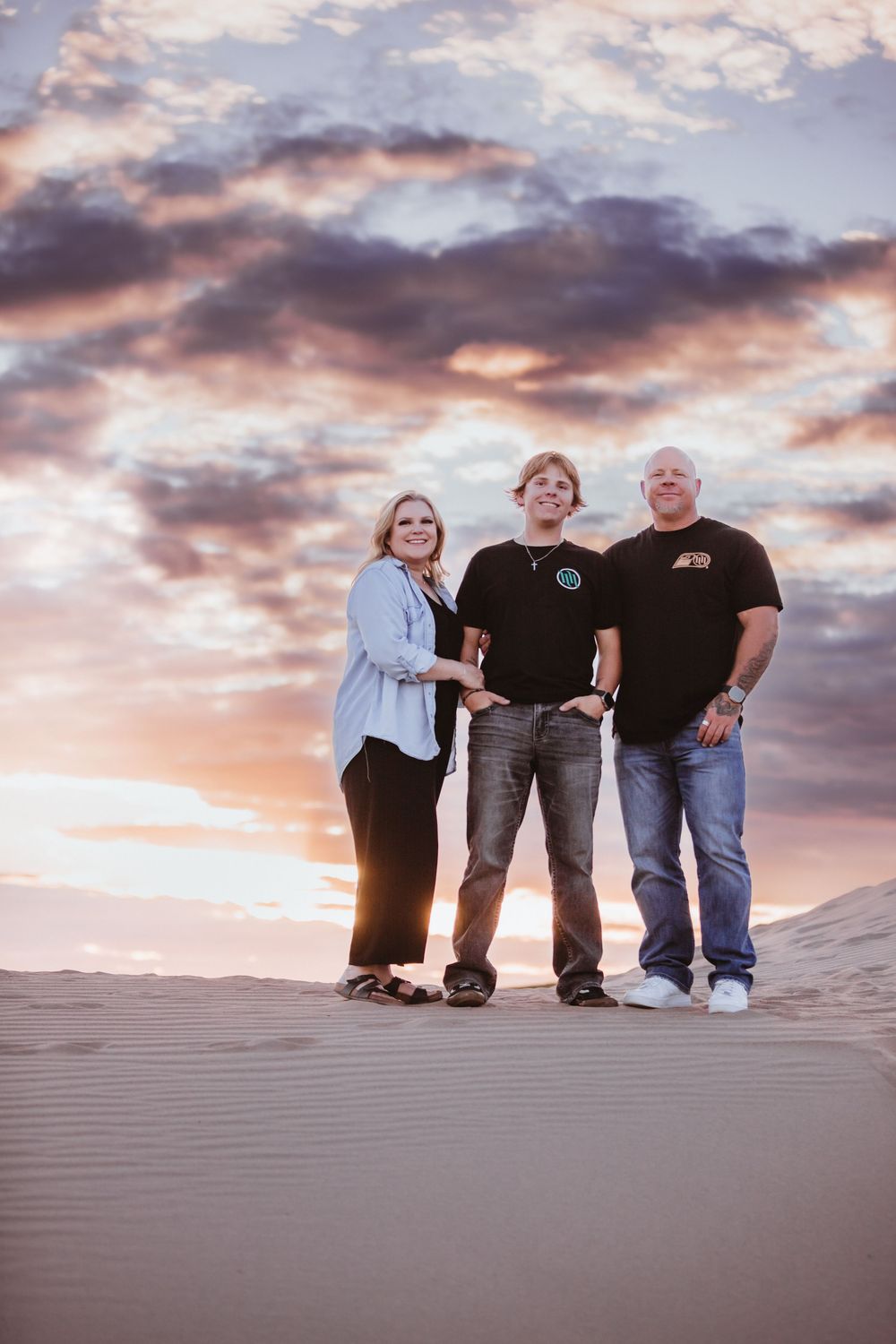 A group of people sit together on sand dunes against a dramatic purple and orange sunset sky.