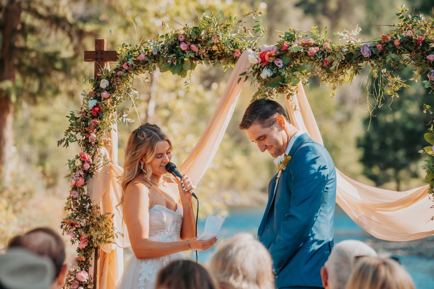 A wedding arch decorated with colorful flowers in front of a lake creates a romantic outdoor ceremony setting.