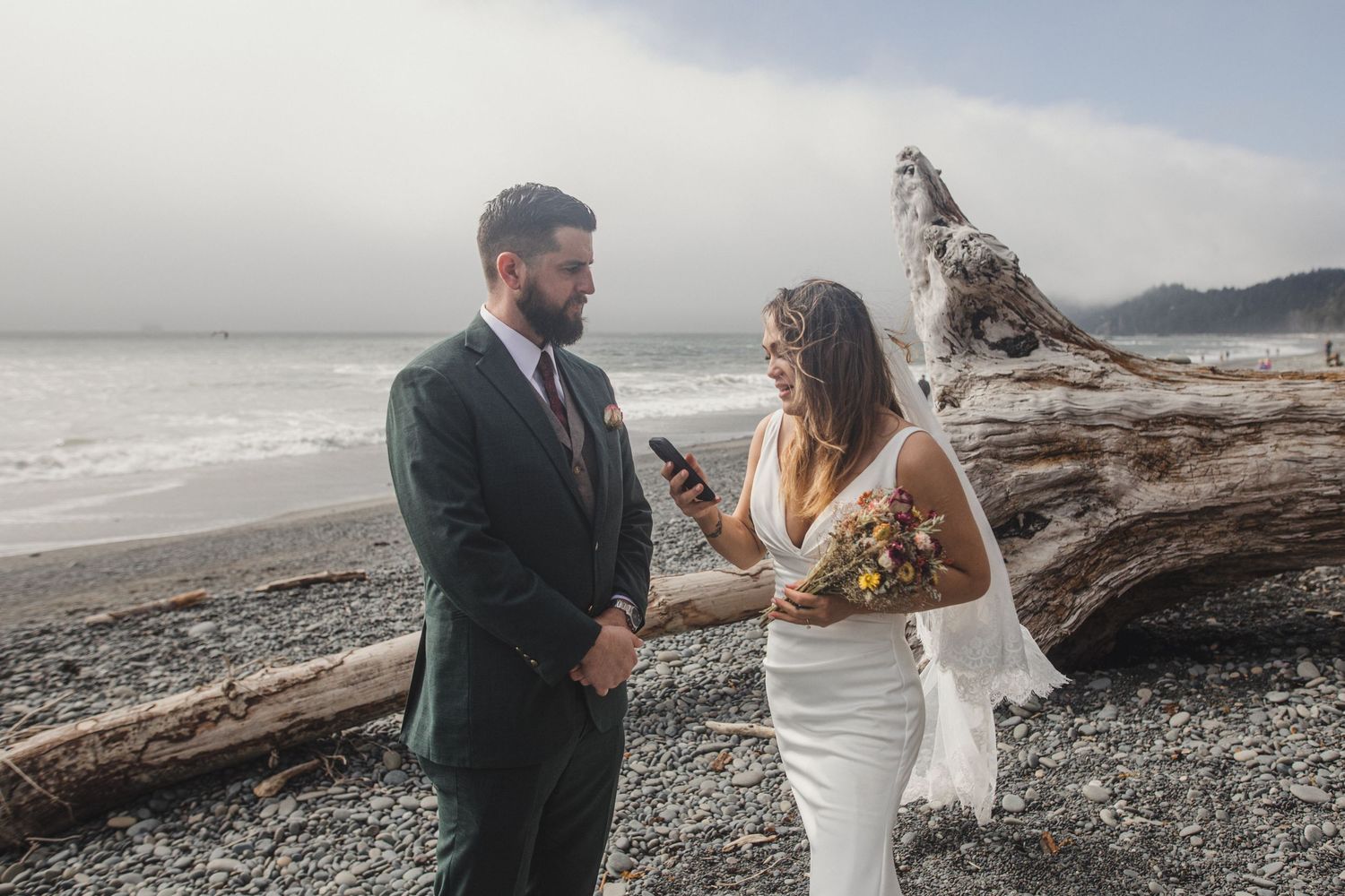 A couple stands together holding hands on a foggy beach with driftwood log and mountains in the background.