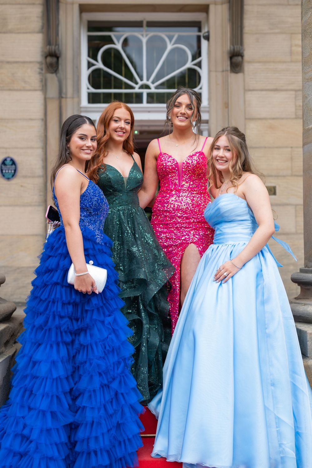 Group wearing formal gowns in blue, green, and pink posing outside a stone building.