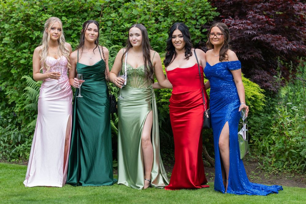 Group wearing formal dresses in pink, green, red and blue poses together in a garden setting.