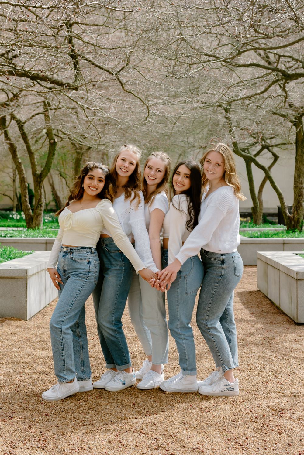 A graduate group sits together on stone steps wearing coordinated white tops and blue jeans in a springtime outdoor setting.