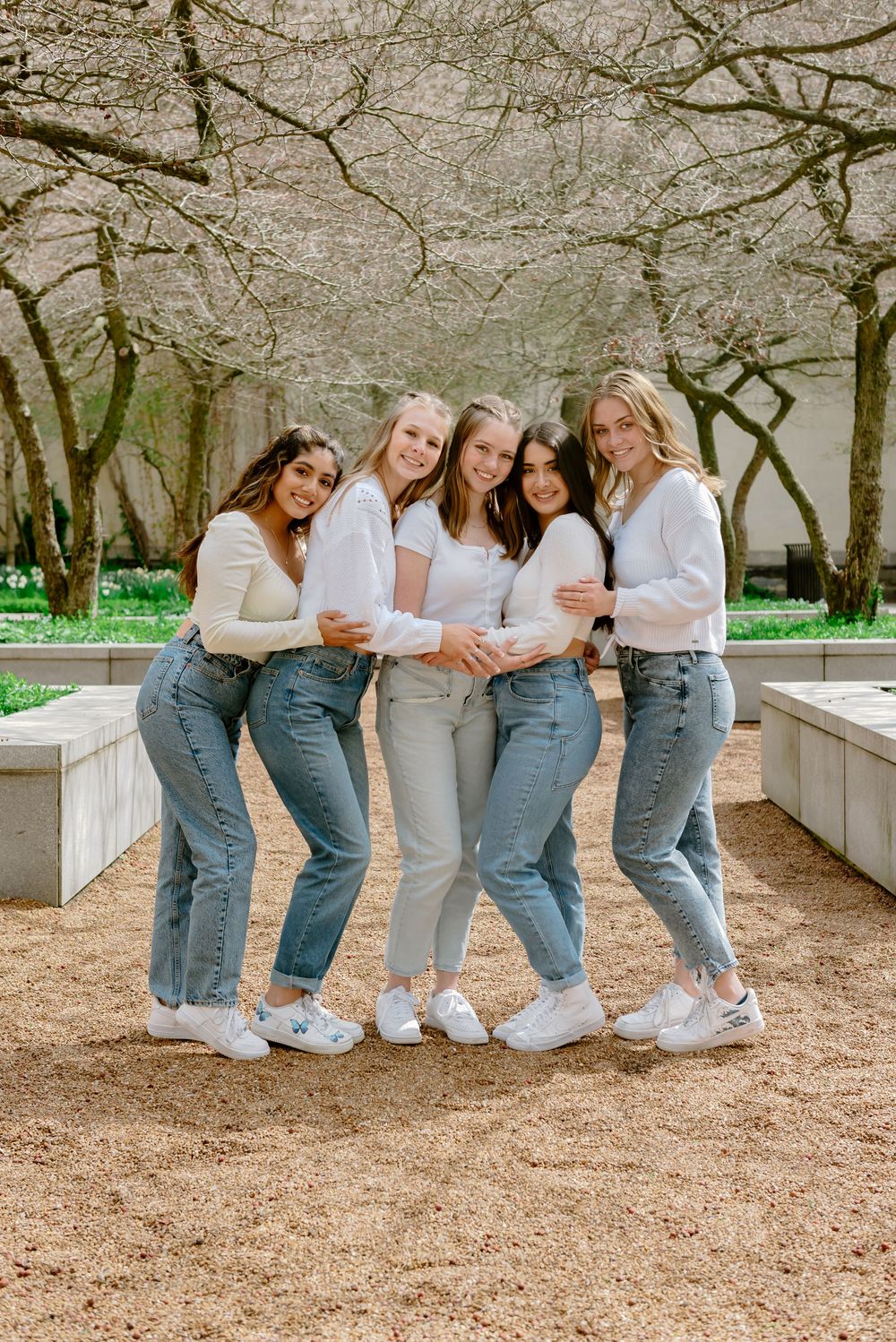 A graduate group of friends wearing white tops and denim jeans pose casually outdoors near blooming spring trees.
