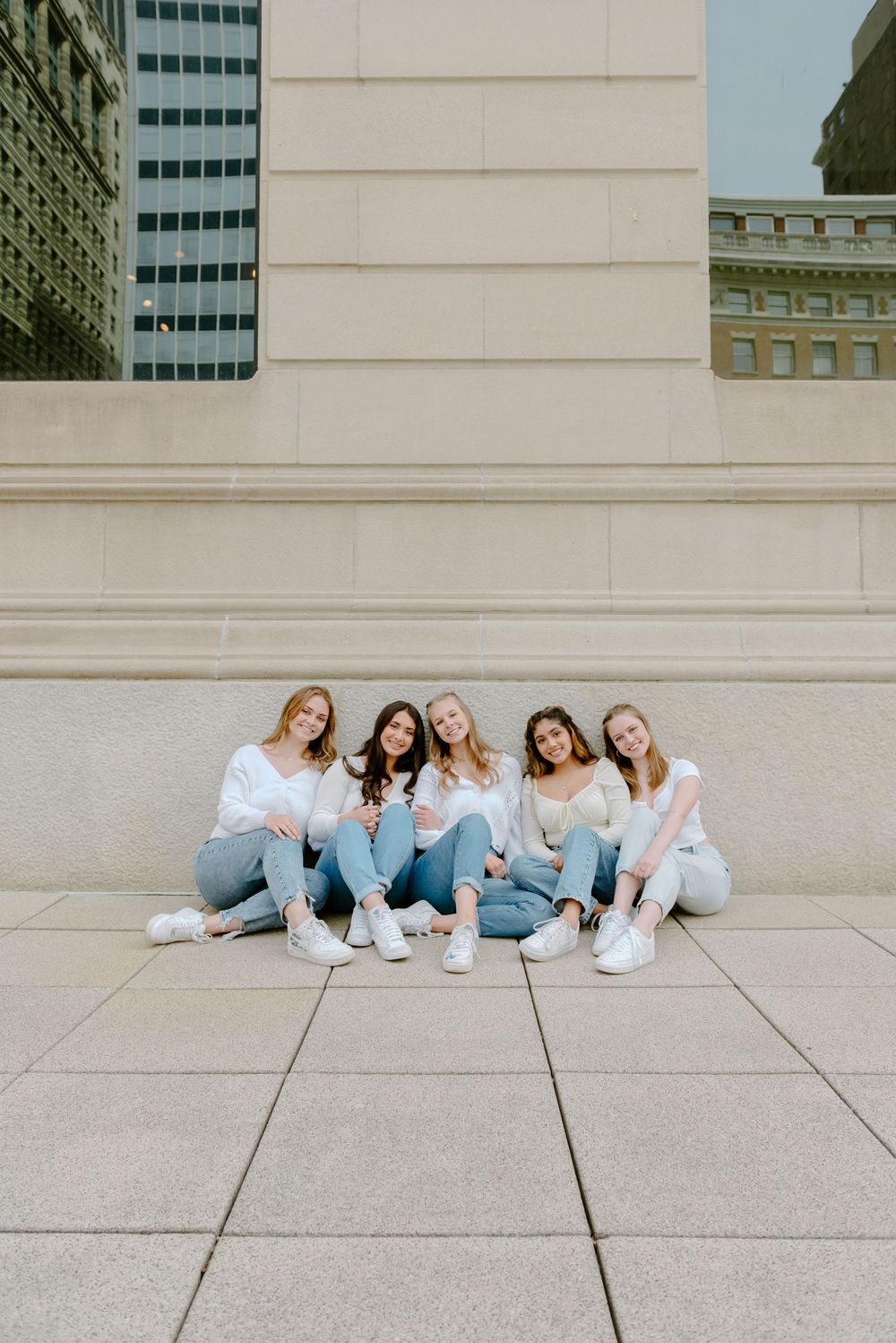 A graduate group in coordinated casual outfits of jeans and white tops sit together on concrete steps of an urban building.