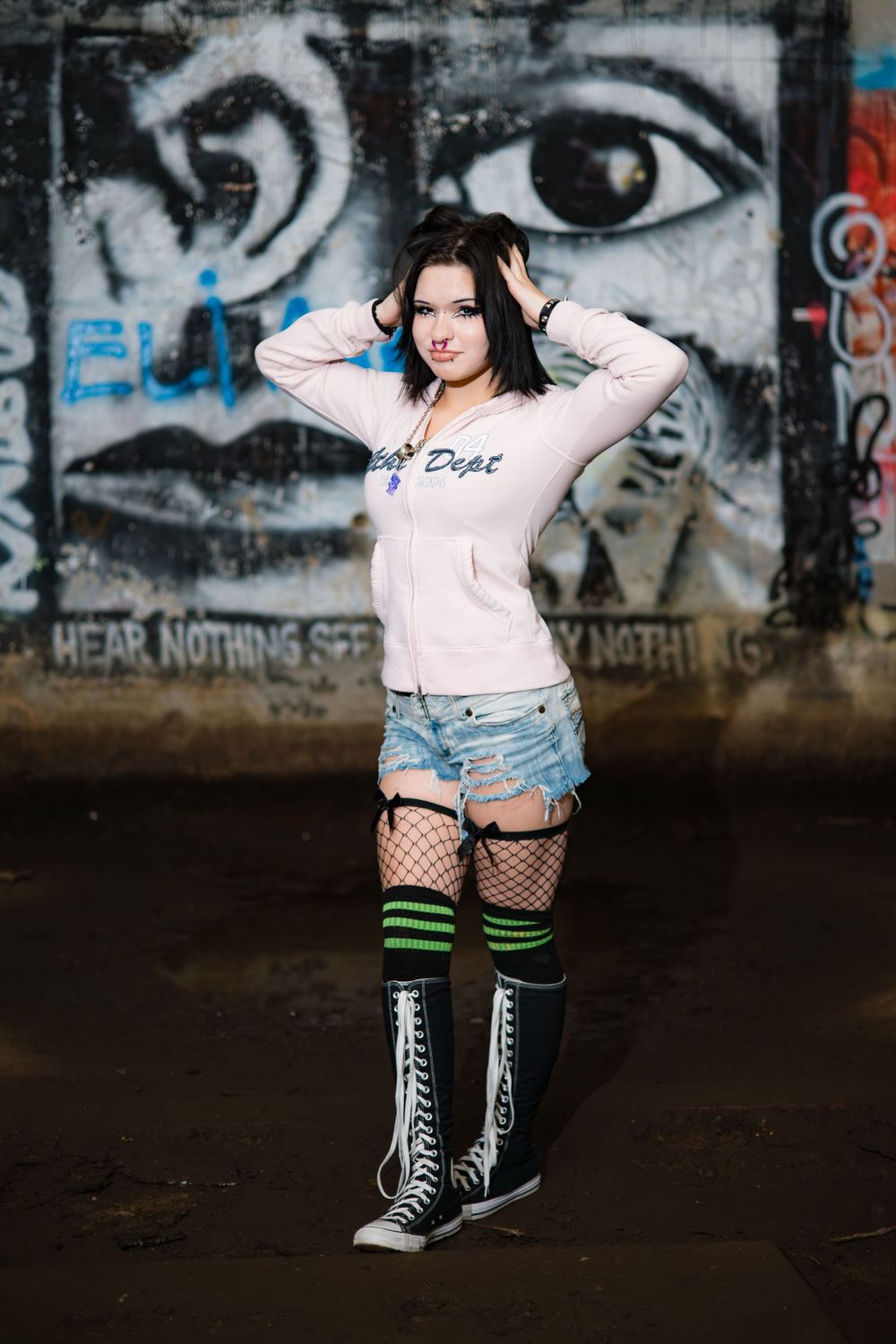 Alternative fashion model poses against graffiti wall wearing fishnet stockings, denim shorts and high top sneakers.