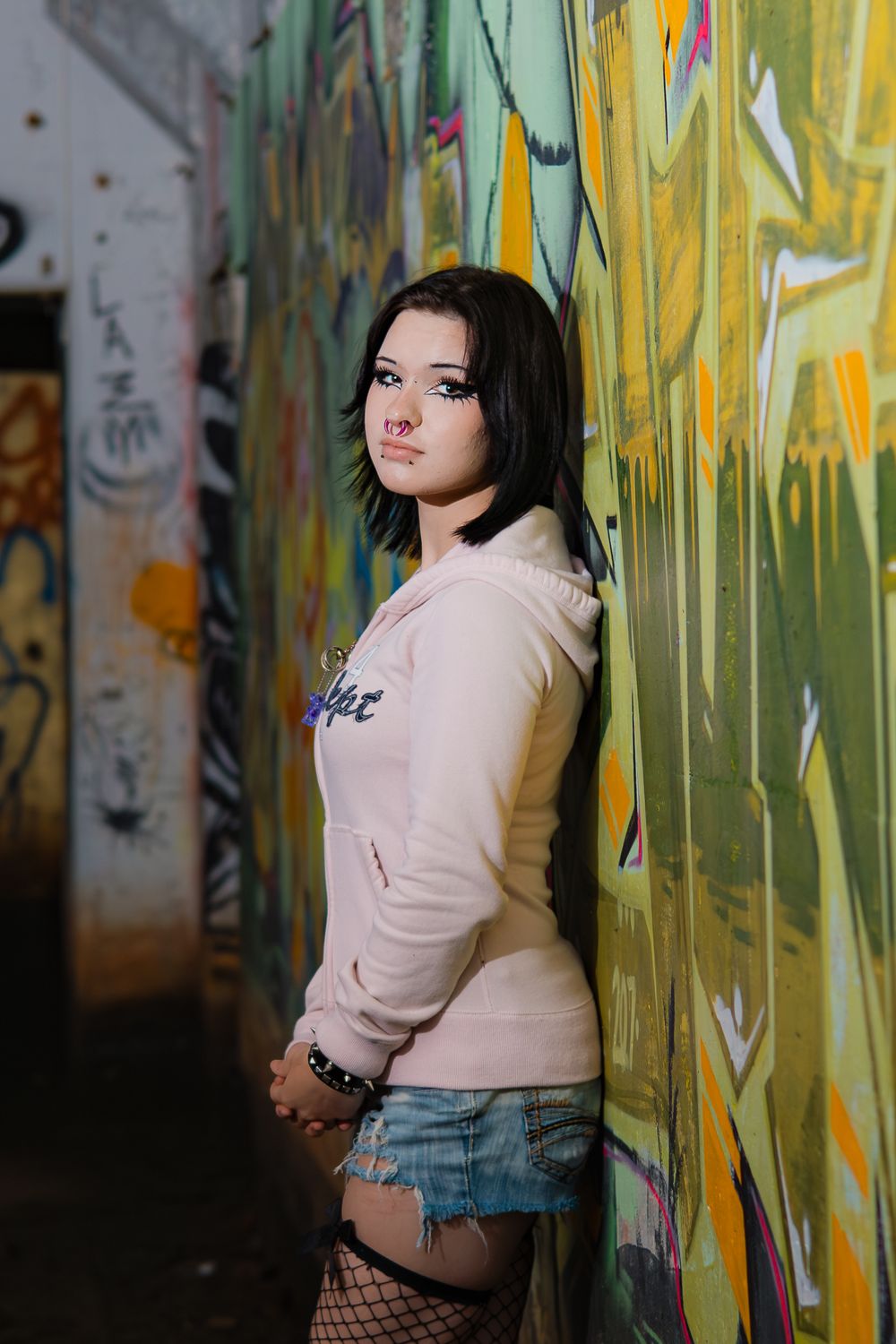 Person wearing pink sweater and denim shorts poses in front of colorful graffiti-covered wall in urban setting.