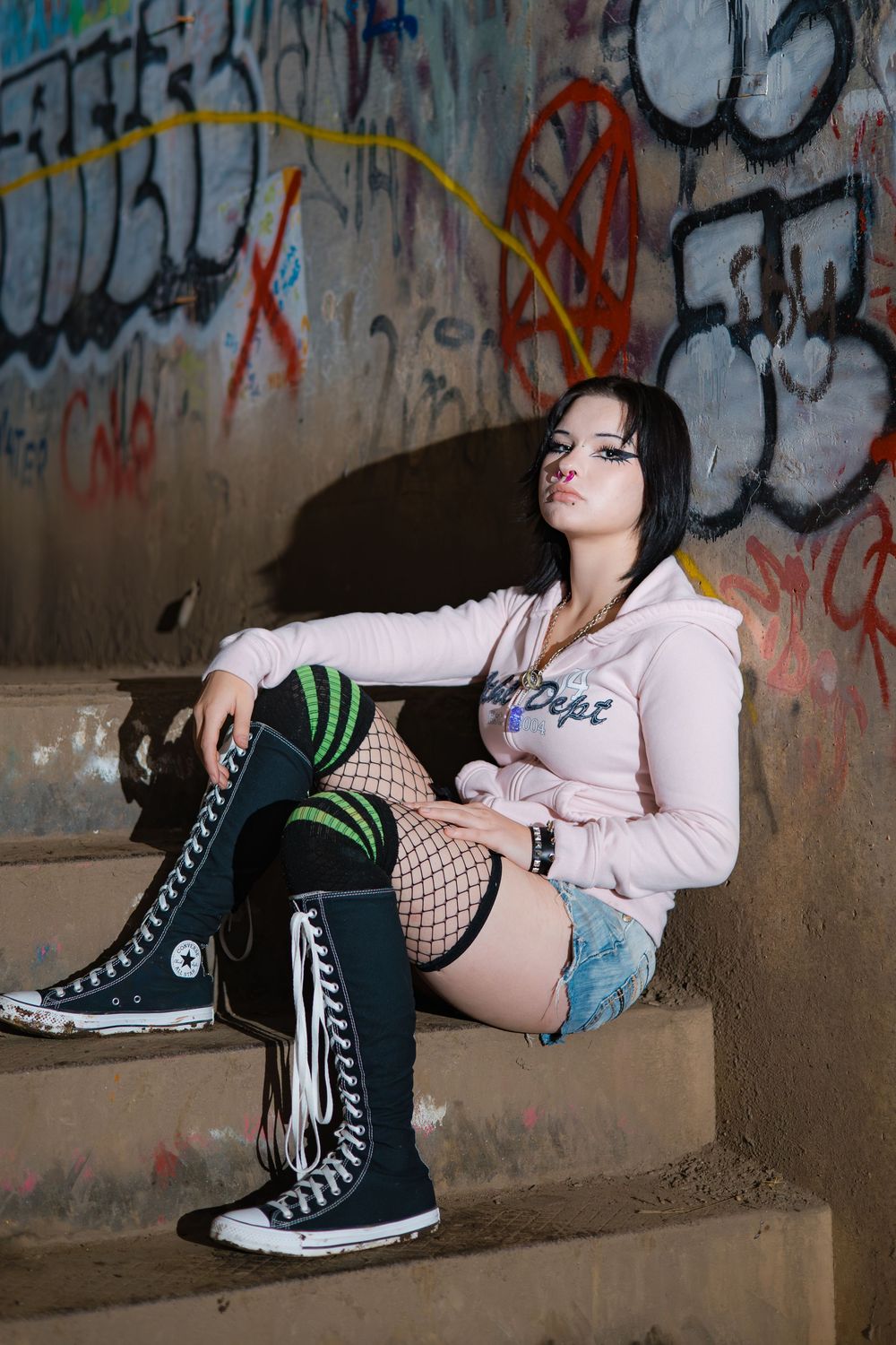 Person in fishnet stockings and black converse shoes lounging against graffiti wall in urban setting.