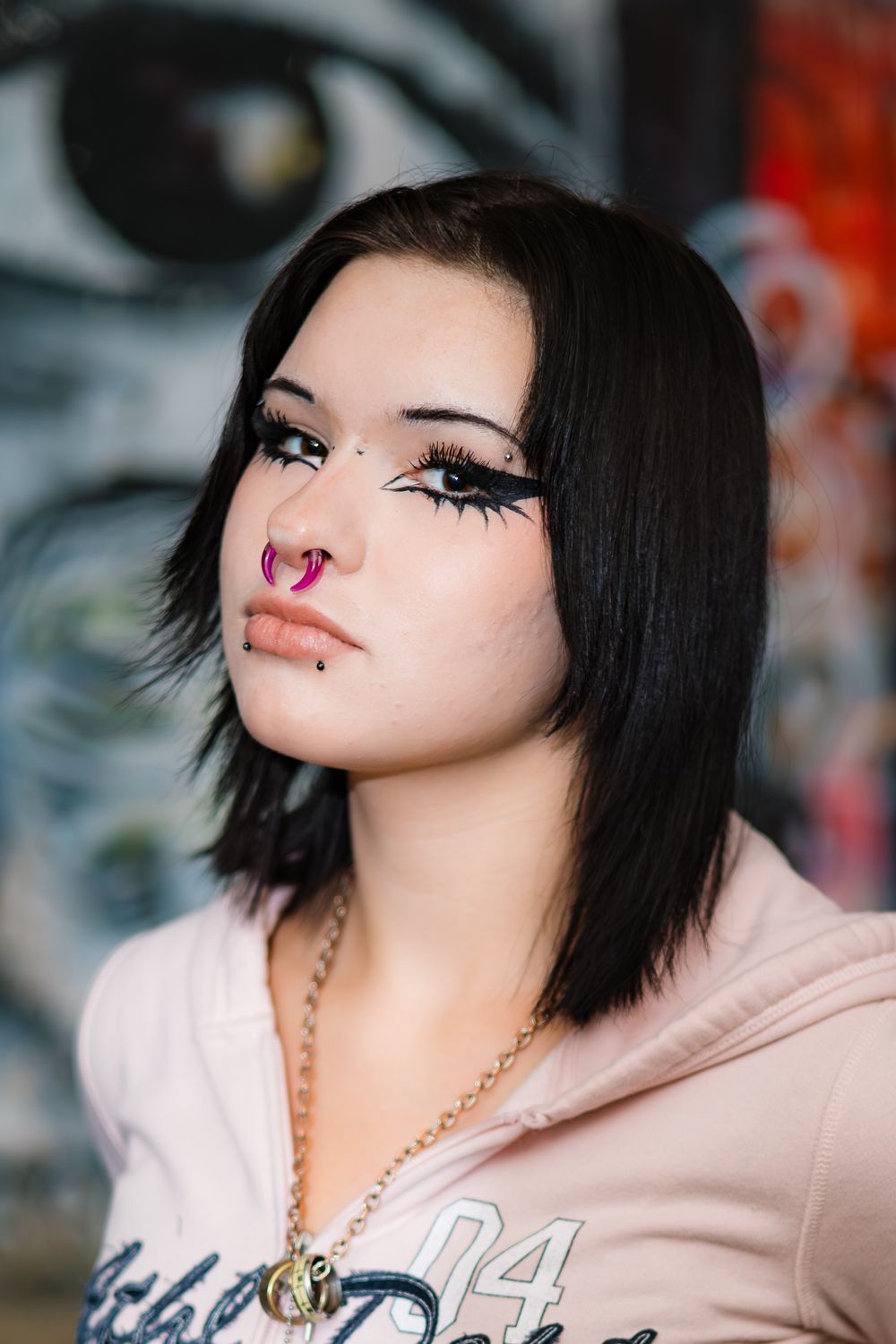 Close-up portrait with black hair, nose piercing and bold makeup wearing a light colored top against a blurred background.