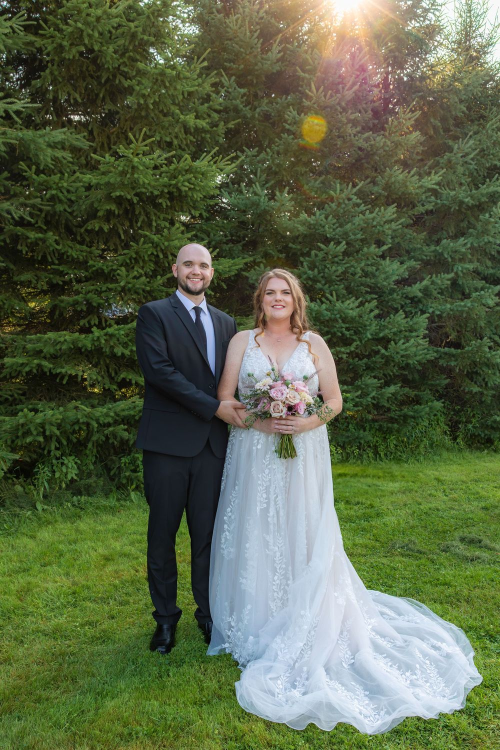 A couple poses outdoors for wedding photos at sunset with trees and greenery in the background.