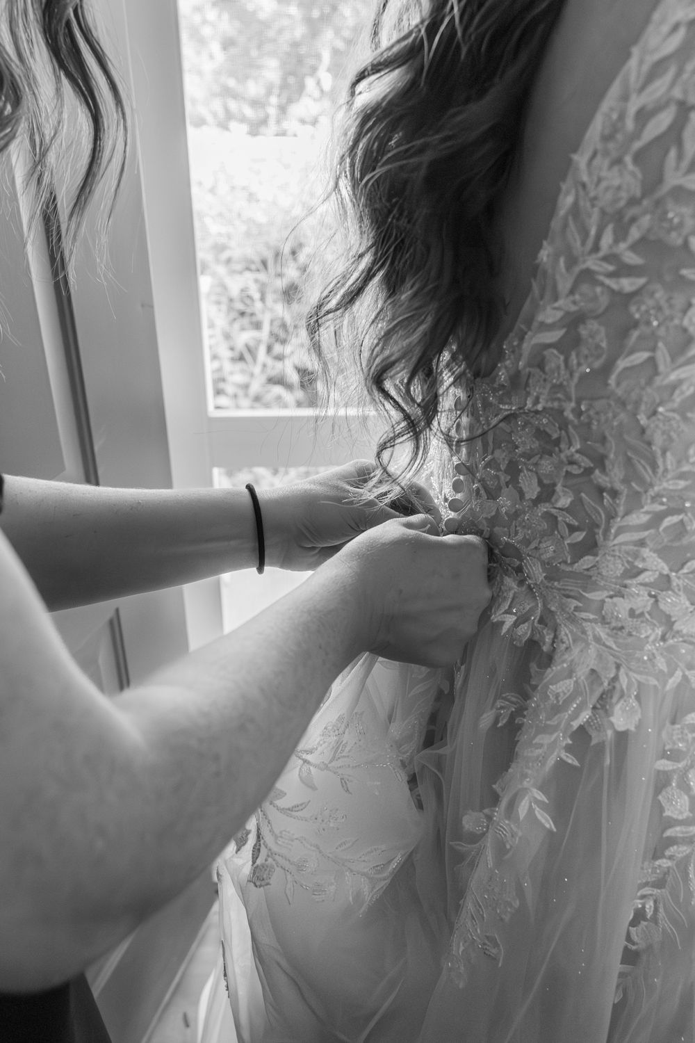 A close-up black and white photo of hands buttoning up an intricate lace wedding dress.