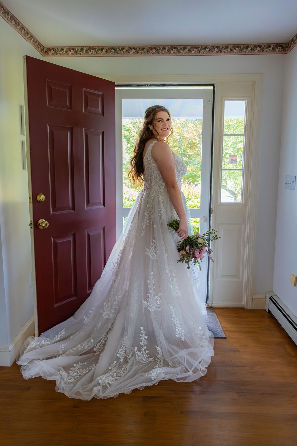 A bride in an elegant lace wedding gown with a long train poses against warm hardwood floors in a sunlit room.