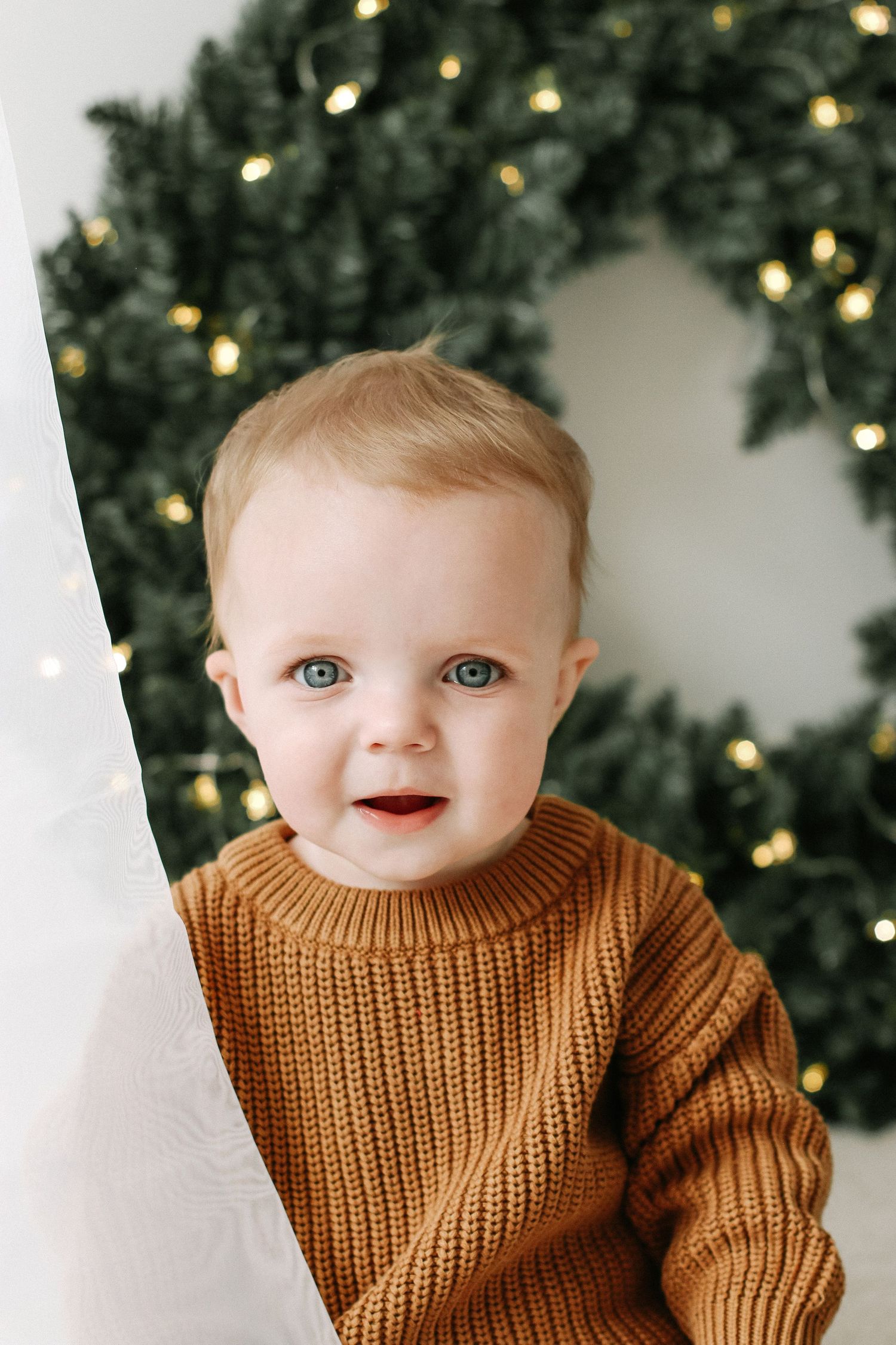 A baby in a cozy brown knit jumper sits in front of a Christmas wreath with twinkling lights in the background during photoshoot Nottingham