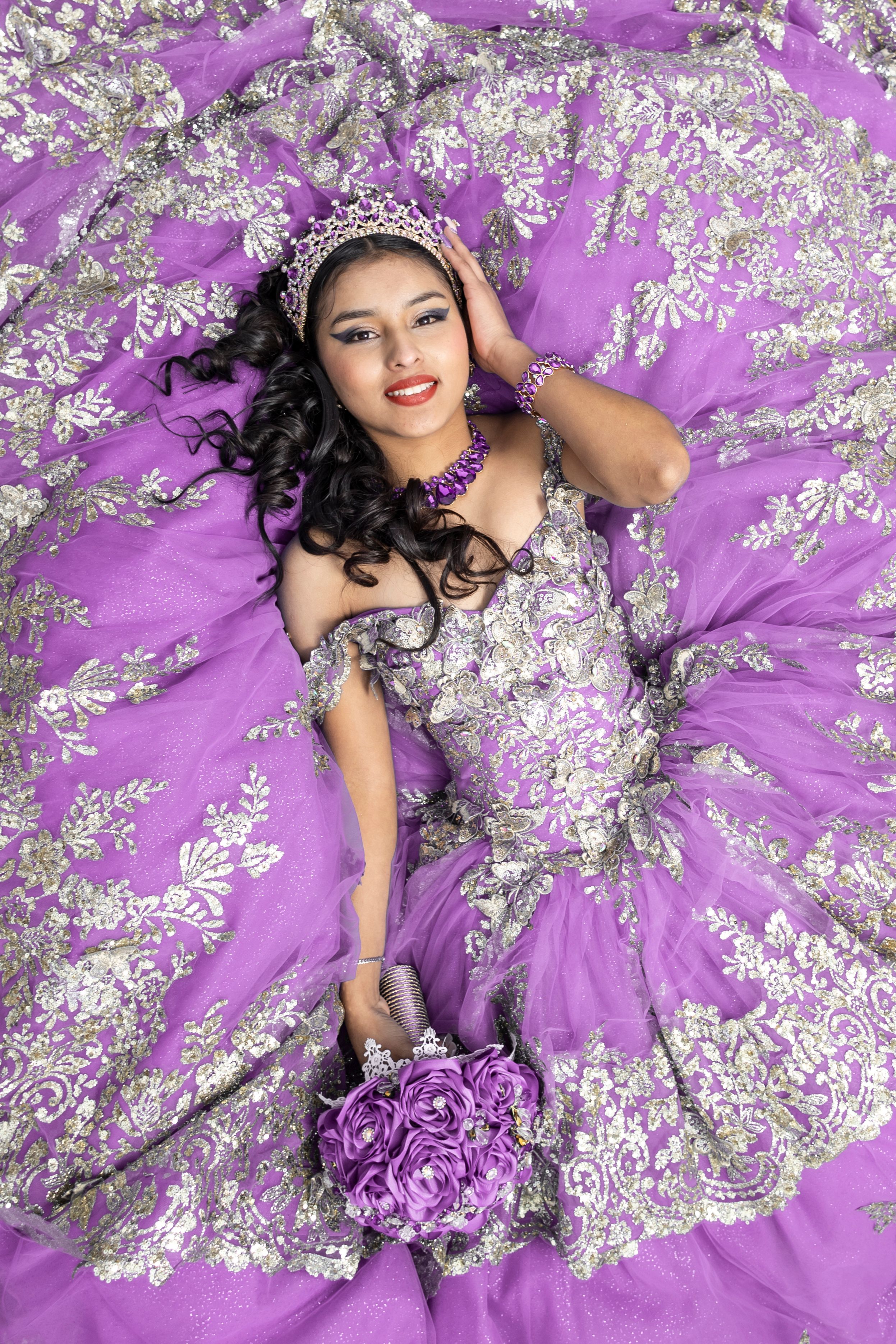 A model in a flowing purple gown poses with white floral decorations for a glamorous fashion photoshoot.