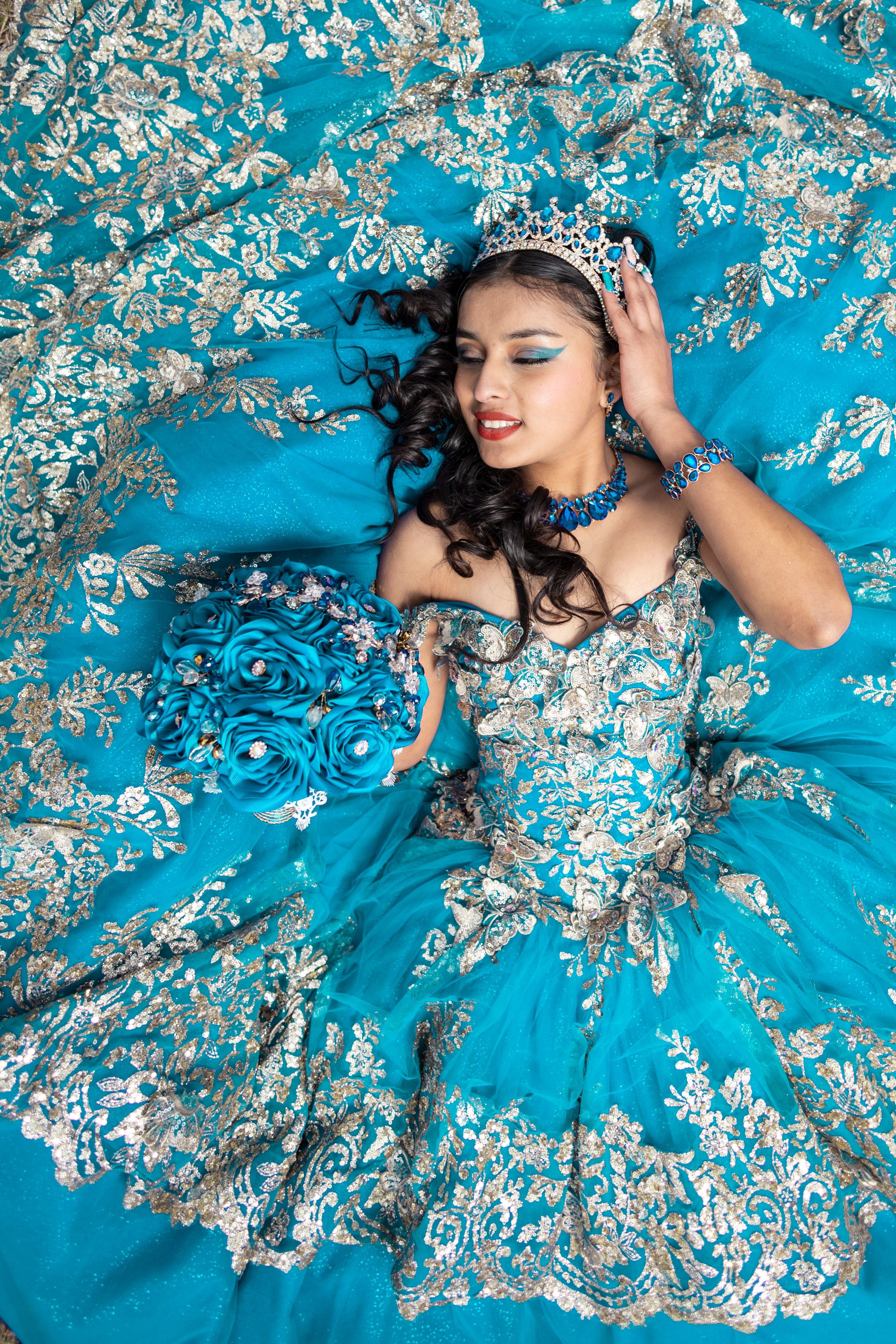 Elegant turquoise quinceañera dress with silver embellishments spreads across the floor in a dreamy ballgown silhouette.