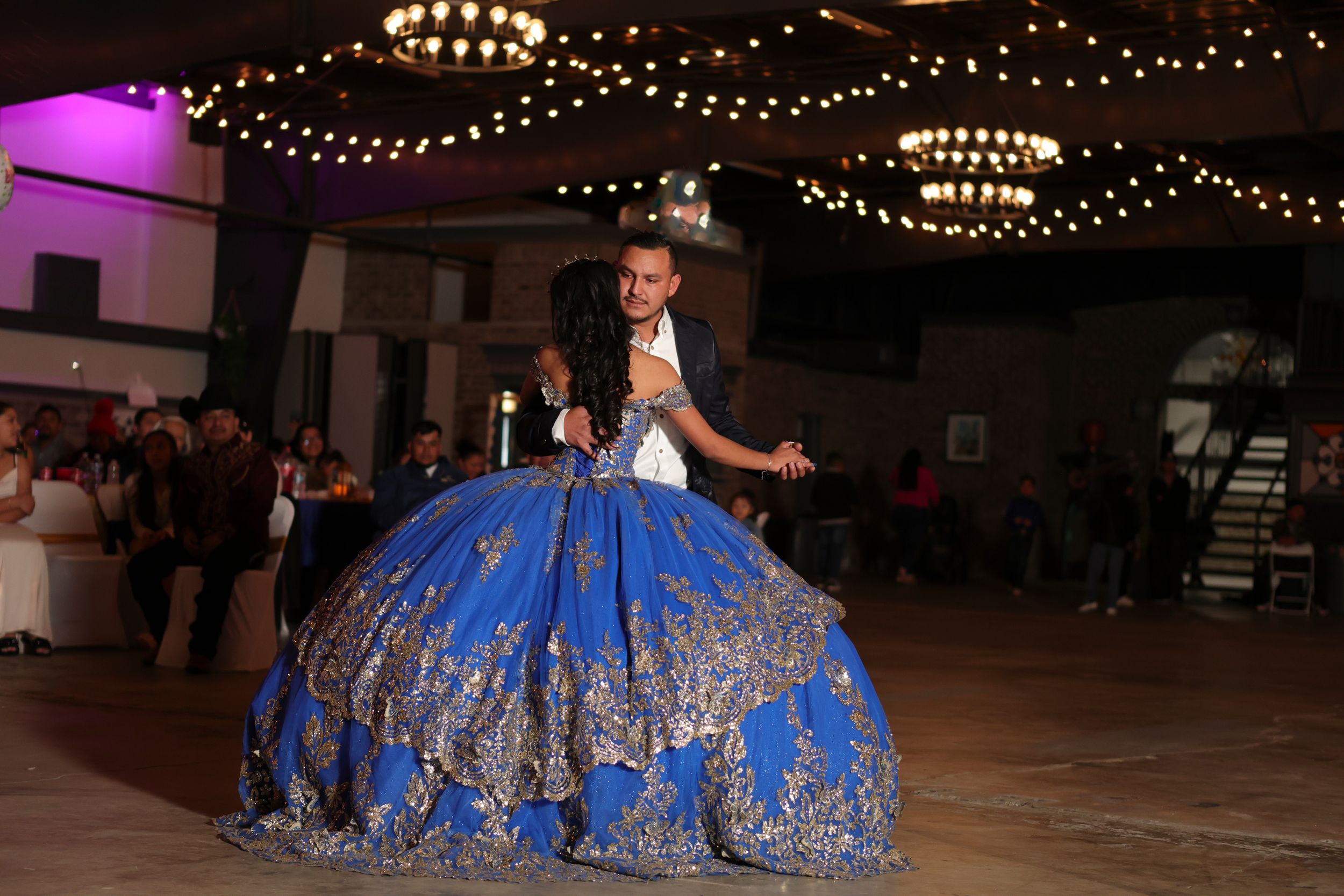 A performer in a flowing royal blue and gold ballgown twirls on a dance floor under warm lighting.