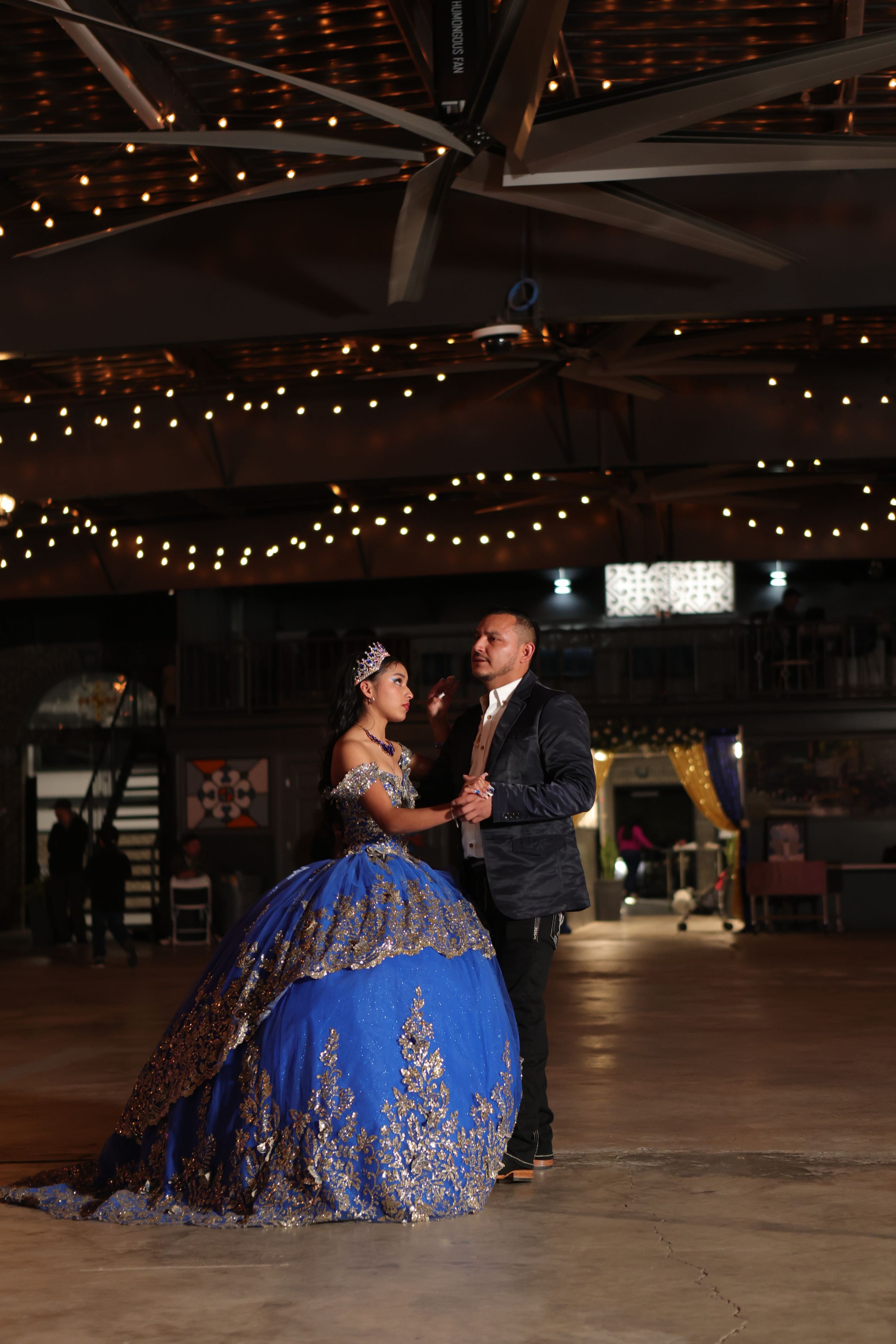 Dancers in formal attire perform on a wooden dance floor under twinkling string lights in a spacious venue.