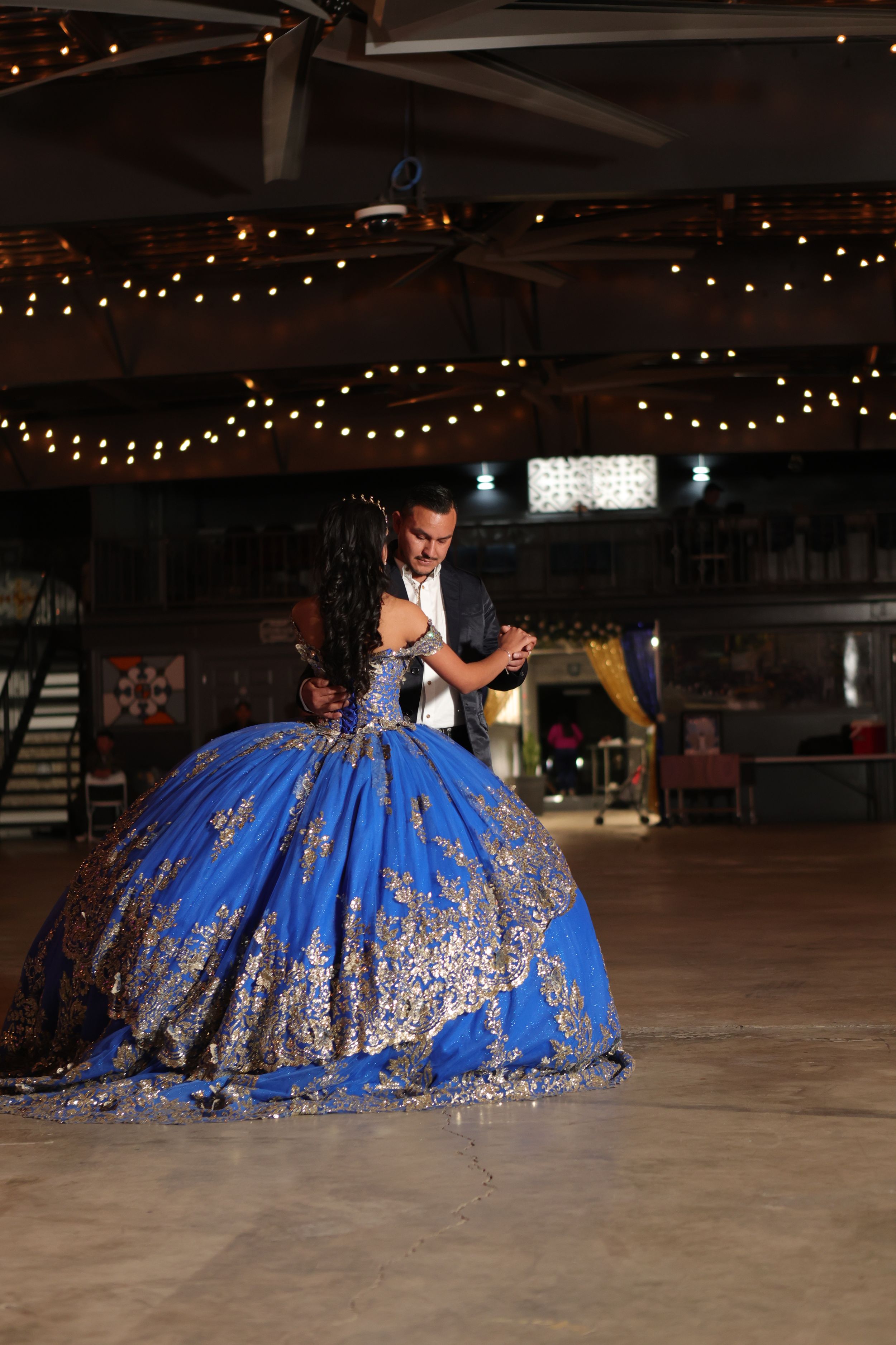 A blue and silver quinceañera dress with sequins sparkles under string lights in an empty indoor venue.