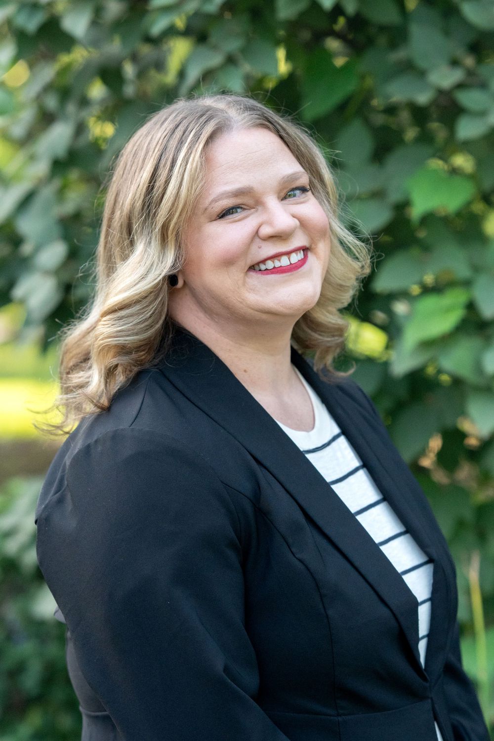Professional woman in black blazer smiling outdoors with green ivy background, blonde hair and red lipstick.