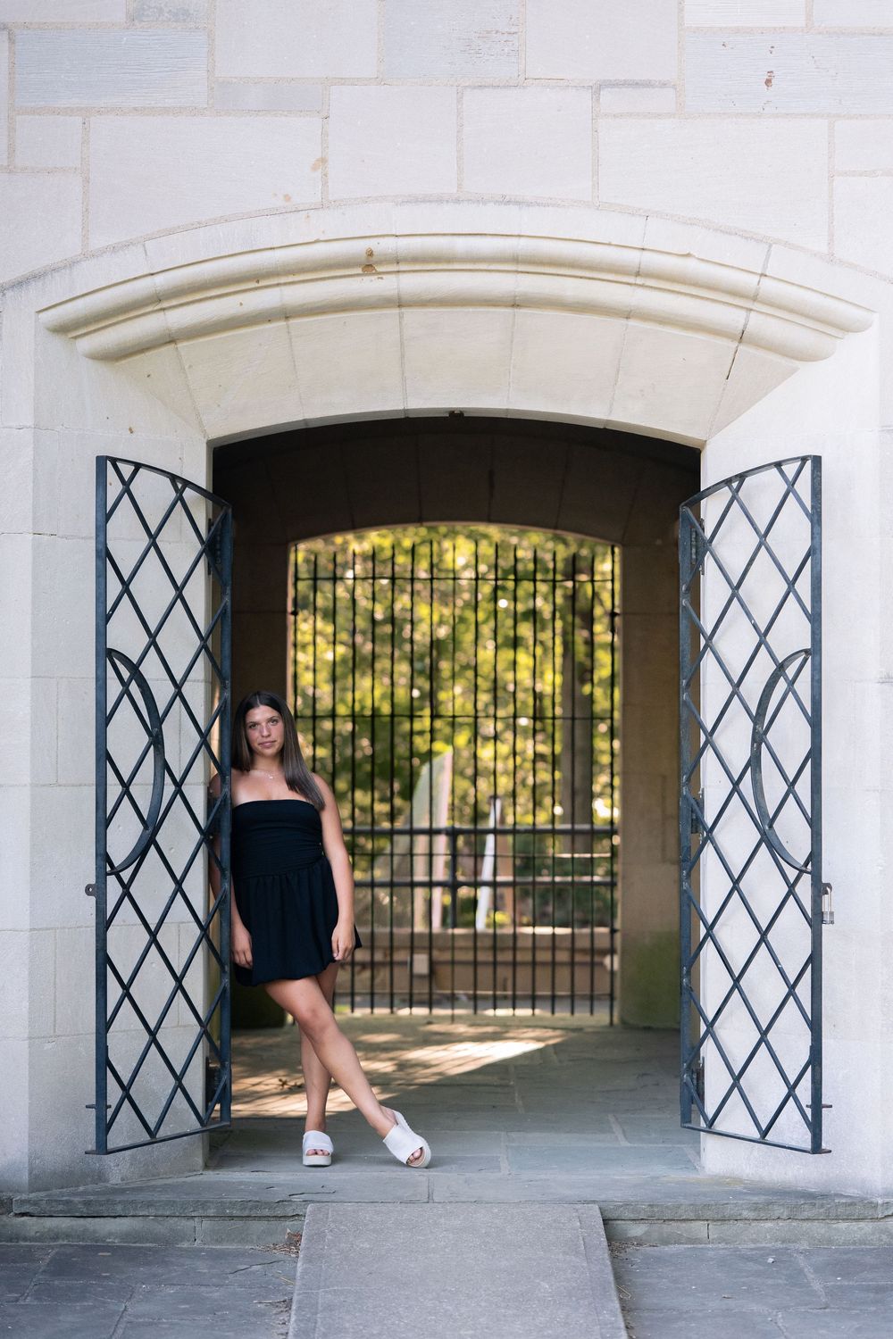 Young woman in black dress poses in ornate stone archway with decorative iron gates and lush greenery behind.