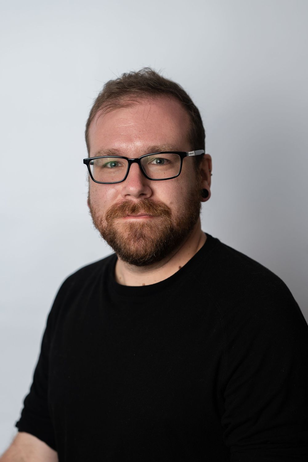 A bearded man with red hair and black rectangular glasses wearing a black t-shirt poses for a professional headshot.