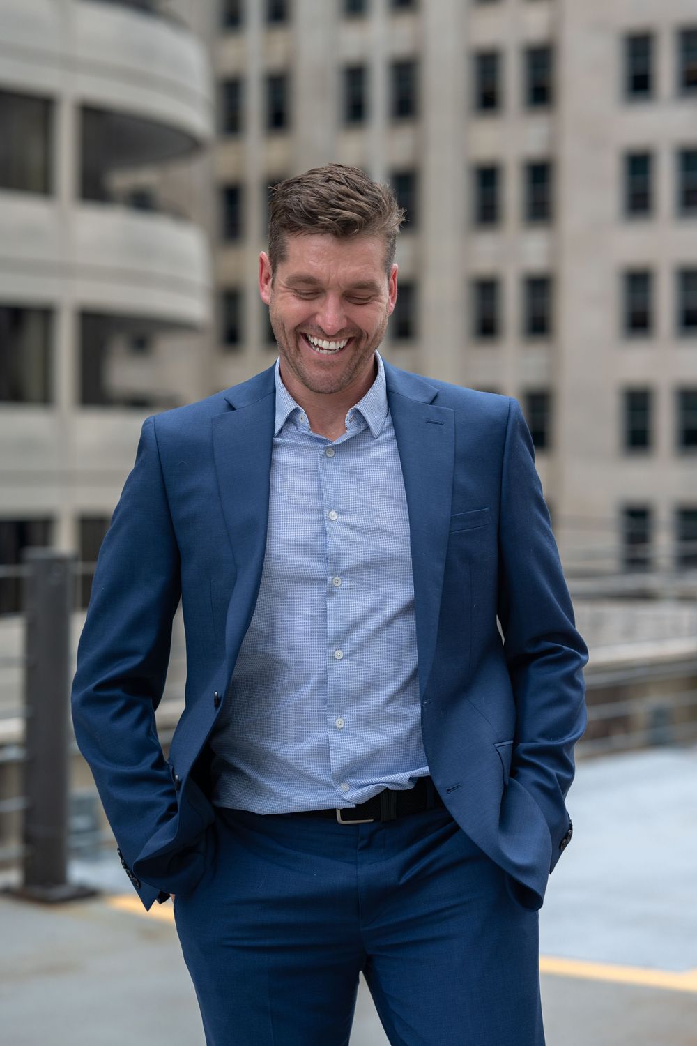 Smiling businessman in navy blue suit standing outdoors with urban buildings in background.